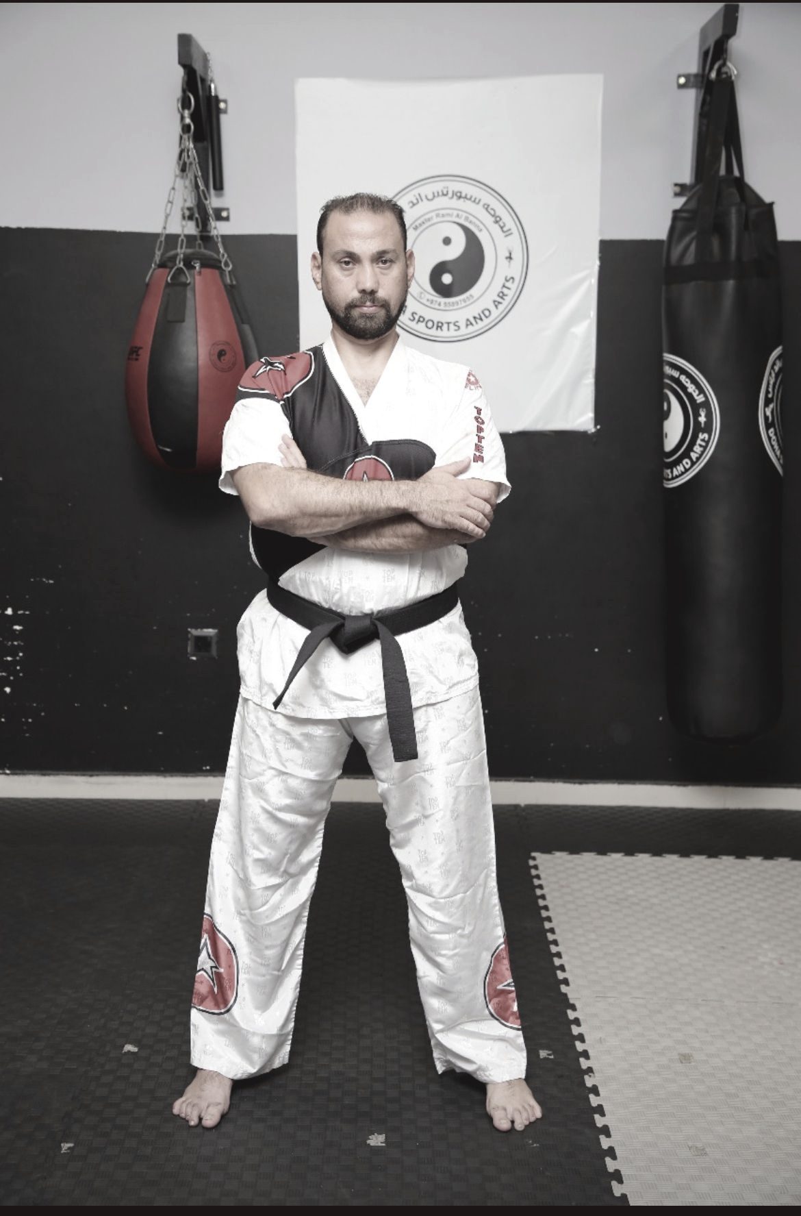 Master Rami Al Banna standing with arms crossed in a white gi and black belt, punching bags hanging either side, the club's stamp on a banner behind him.