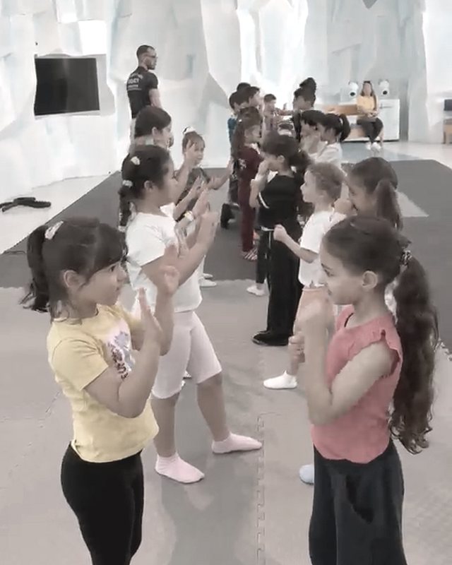 A kids' class on grey mats in front of a blue and white ice-cave backdrop, the children working through drills in pairs while a coach stands at the back.